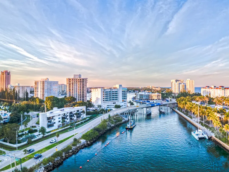 Coastal cityscape with a river, bridge, and buildings under a partly cloudy sky.