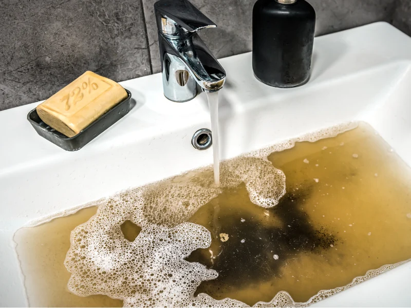 Bathroom sink filled with dirty, brown, soapy water under a running faucet.