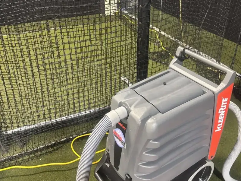 Portable KleenRite cleaning machine with hoses on artificial turf near netting.