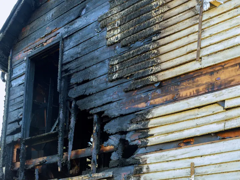 Burned wooden exterior wall of a house with charred and damaged siding near a window.