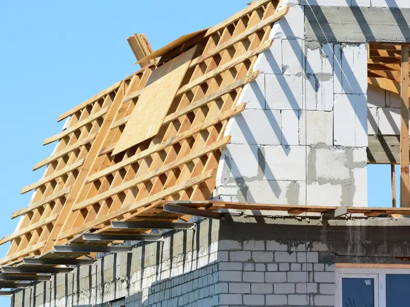 Partially constructed house with wooden roof framework against a clear blue sky.