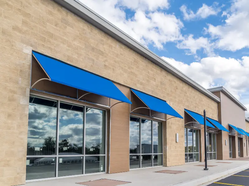 Storefront windows with blue awnings on a beige brick building under a partly cloudy sky.