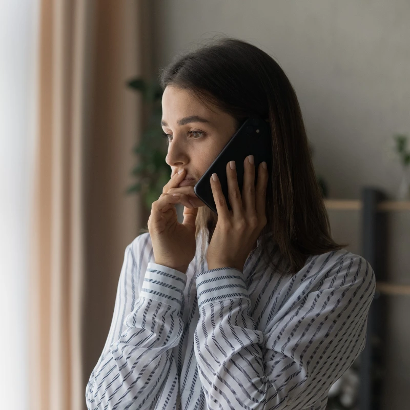Woman in a striped shirt looks thoughtfully out a window while talking on a phone.