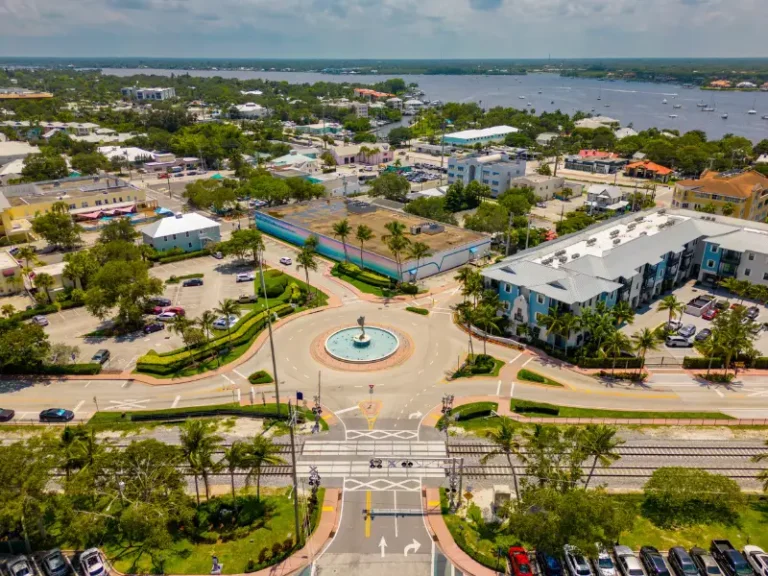 A roundabout with a central fountain surrounded by buildings, palm trees, and a nearby waterfront.