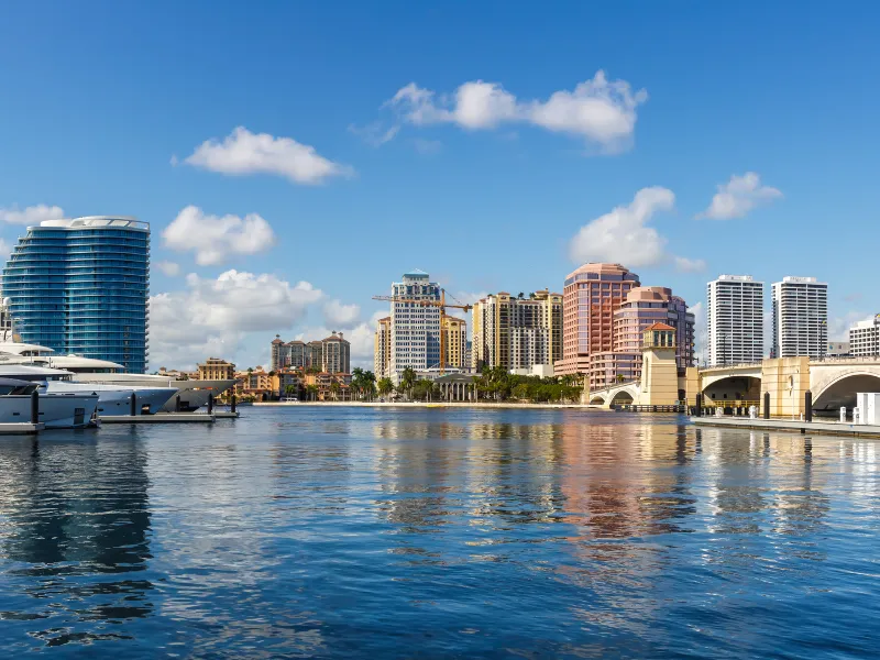 Waterfront city skyline with modern buildings, yachts, and a bridge under a blue sky.